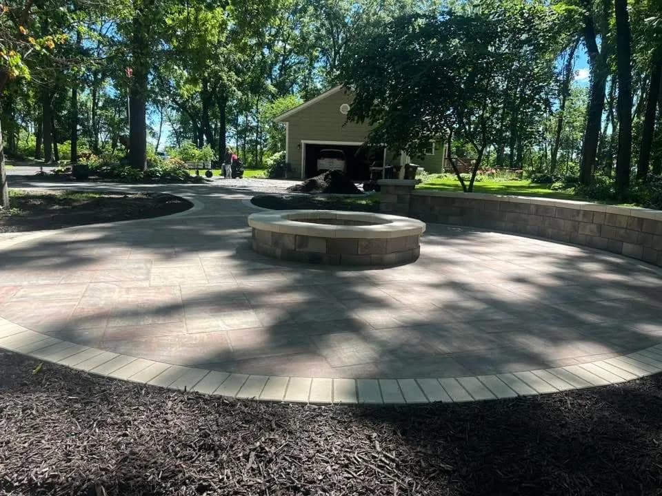 Circular paver patio with a round stone fire pit and a contrasting border ring set in a wooded yard.