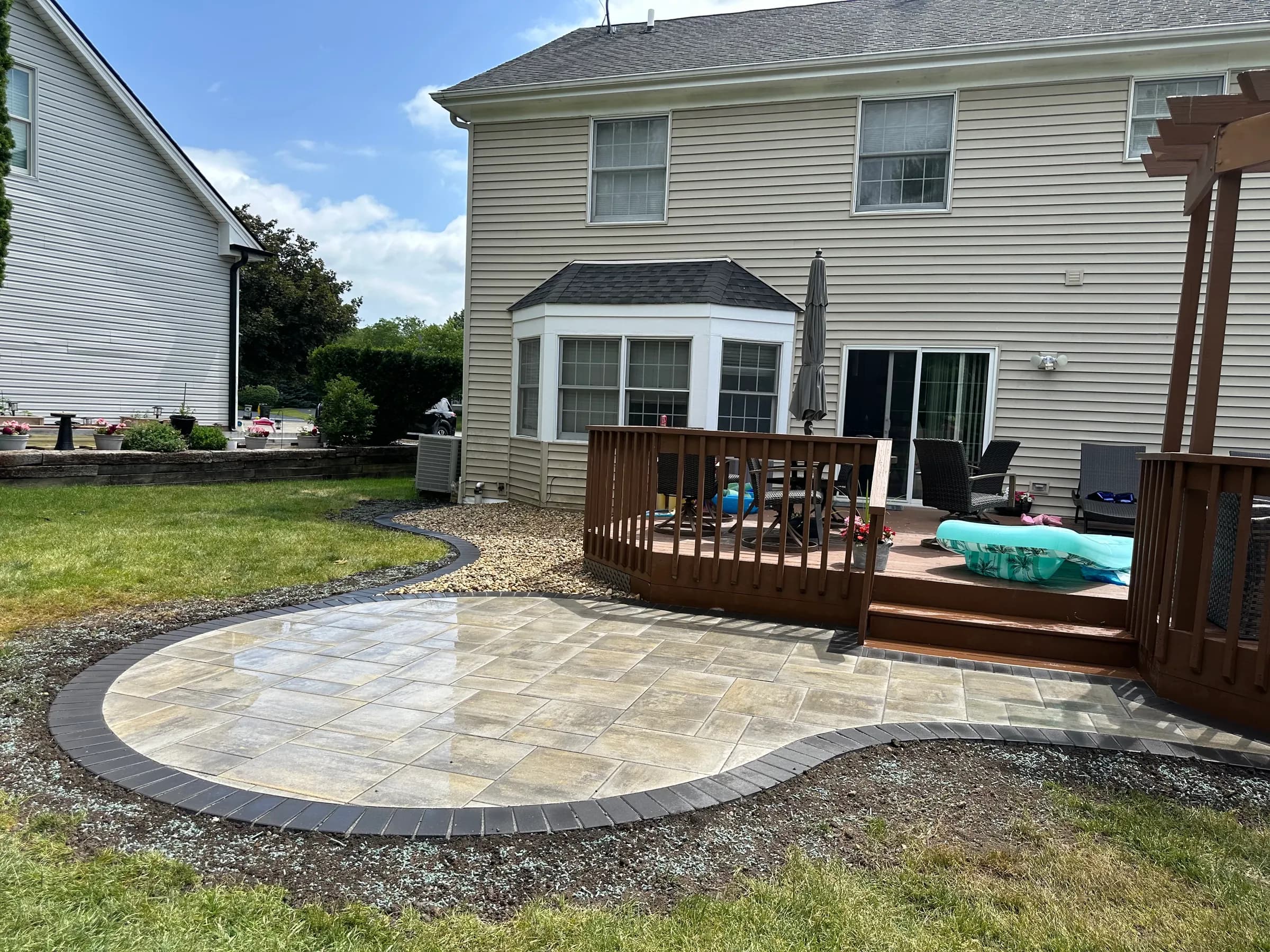 Curved paver patio with a charcoal soldier-course border beside a wood deck, built by E.R. Hardscapes in Northern Illinois.