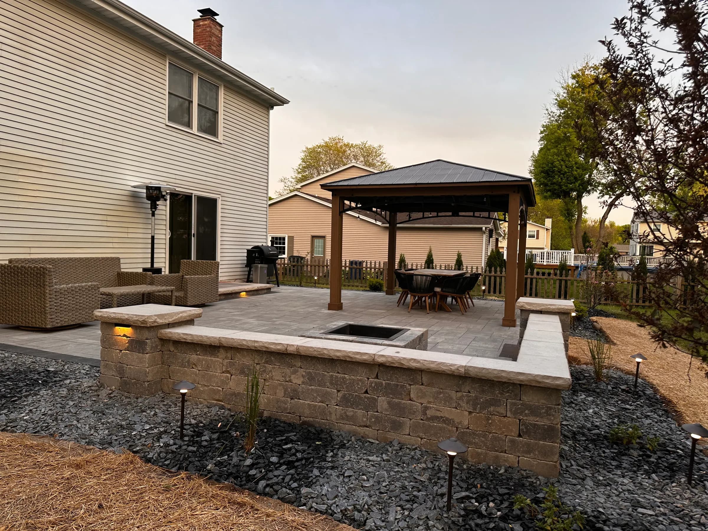 Gray paver patio at dusk with a square fire pit, lit seat wall and stone columns beside a cedar gazebo.