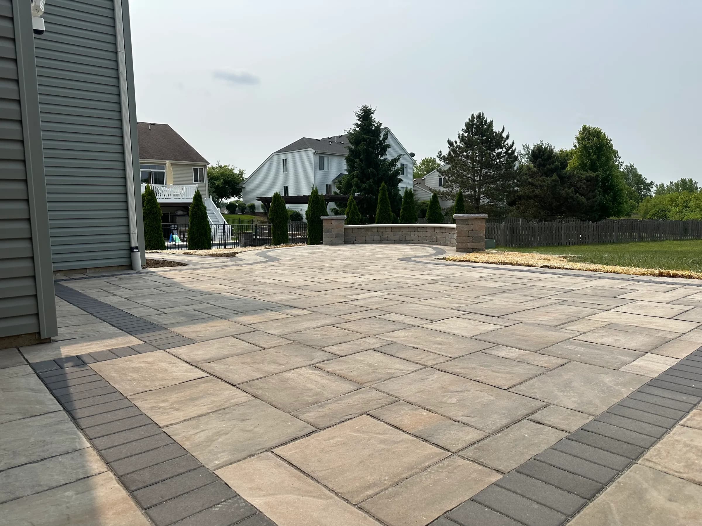 Large tan paver patio with a soldier-course border and a curved seat wall with lit stone columns.