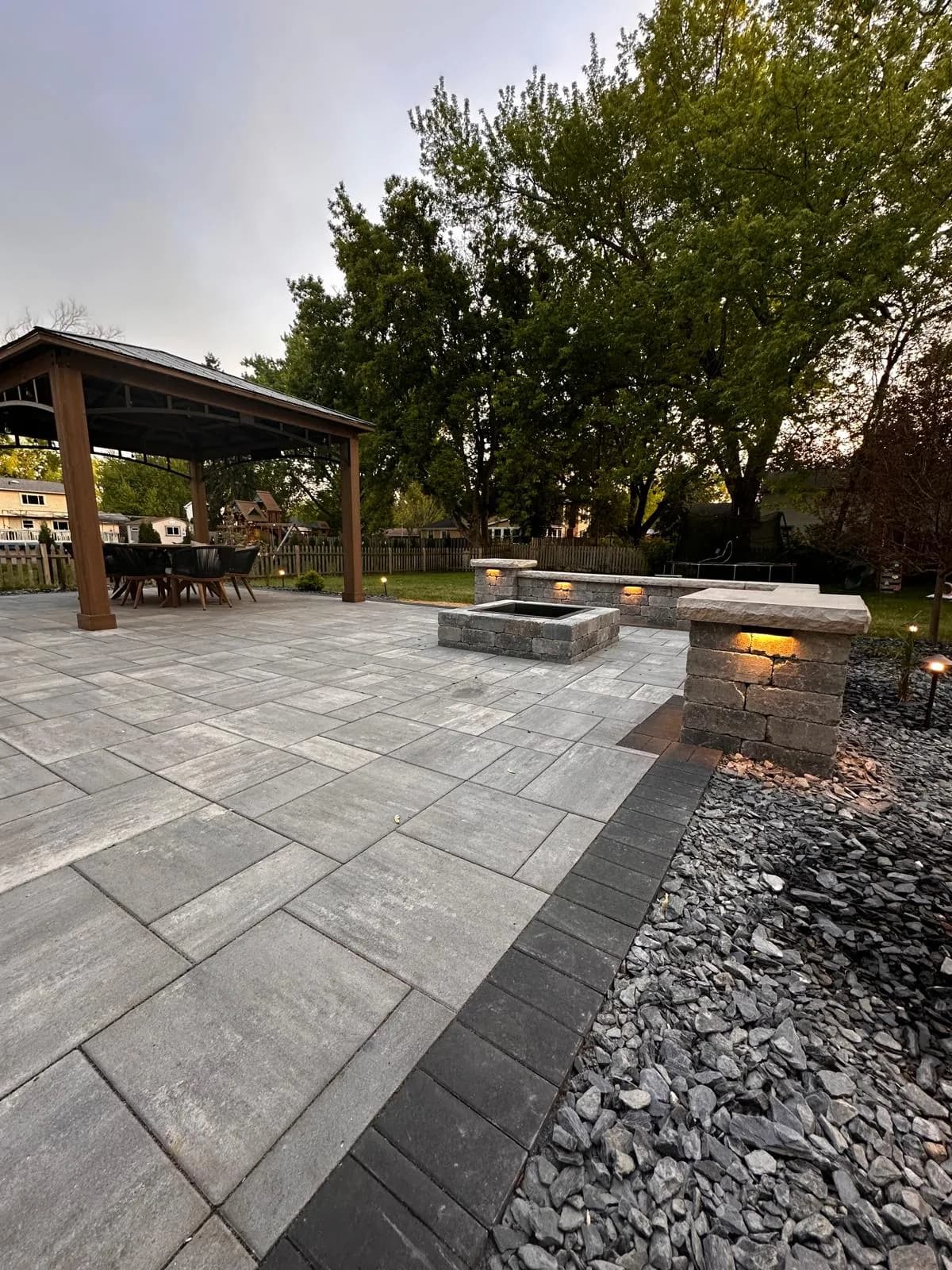 Gray paver patio with a square fire pit and lit stone columns beside a cedar gazebo at dusk.