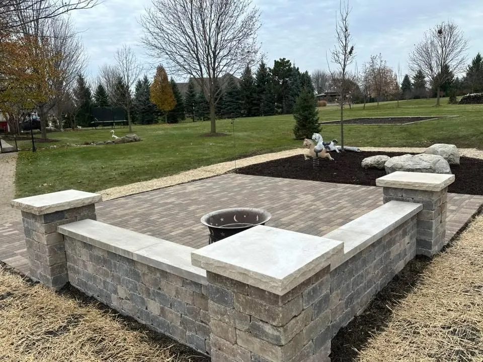 Fire pit with a U-shaped seat wall and capstone columns on a paver patio beside an open lawn and mulch beds.