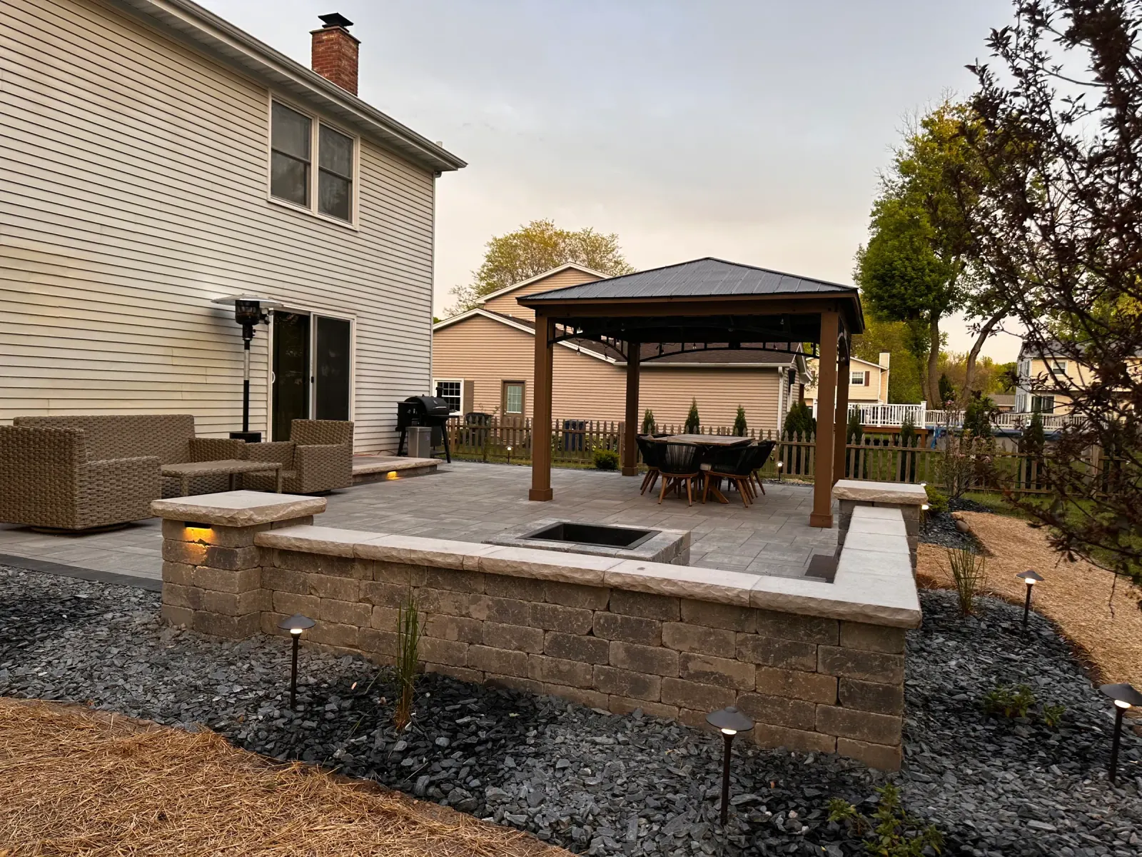 Gray paver patio at dusk with a square fire pit, lit seat wall and stone columns beside a cedar gazebo.