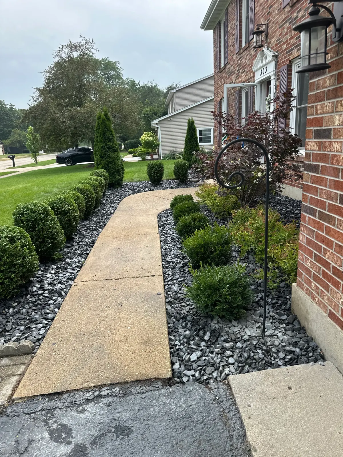 Front-yard landscape beds with decorative rock, trimmed shrubs and a concrete walkway maintained by E.R. Hardscapes.