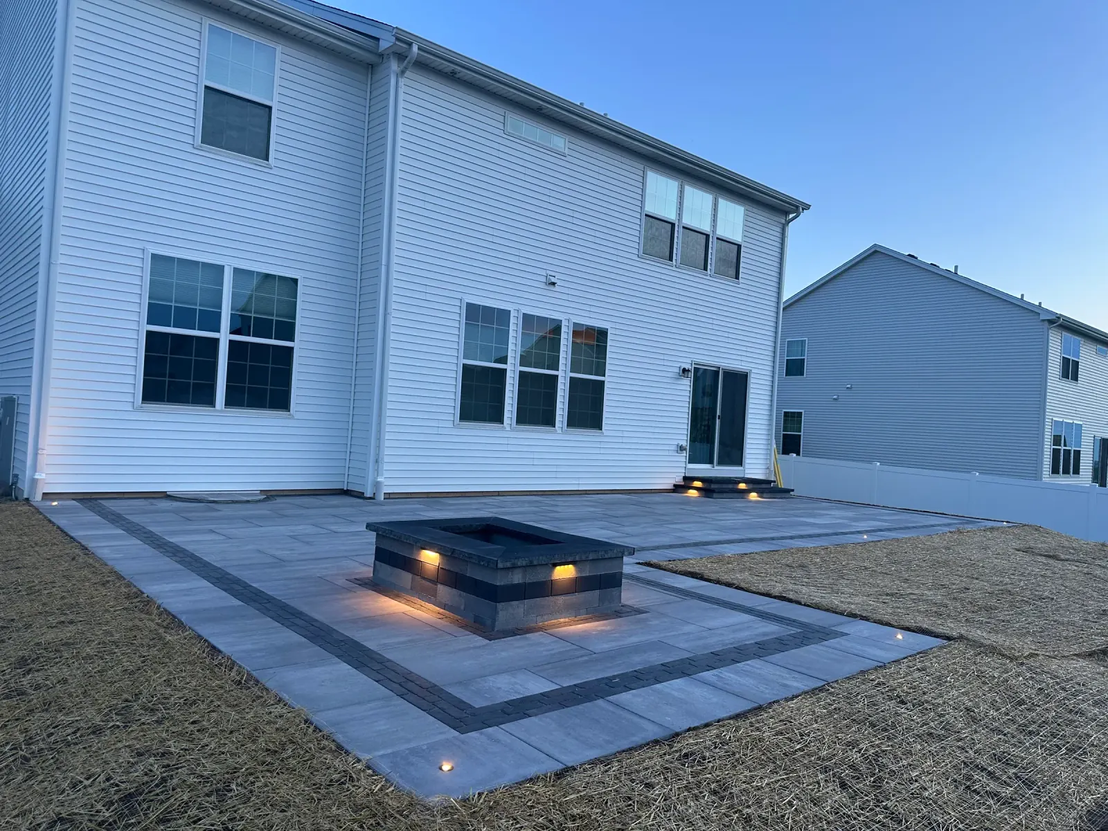 Large paver patio at dusk with a lit rectangular fire pit and symmetrical in-ground landscape lighting.